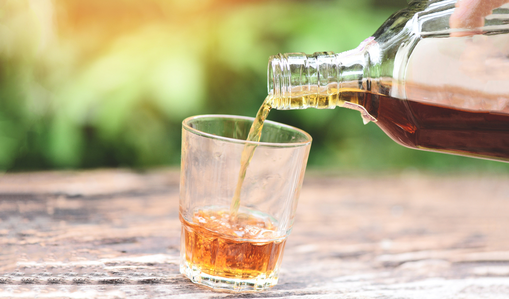 在木质背景上将威士忌或酒精饮料从瓶子倒入玻璃杯倒酒